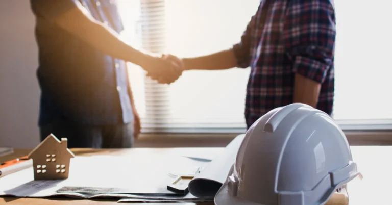 Two people shaking hands in front of a table with a hard hat and documents on top, choosing the right custom home builder.