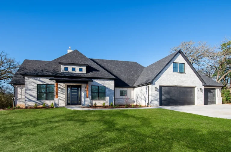 Modern single-story house with gray brick exterior, black roof, and a three-car garage, set against a clear blue sky and green lawn.
