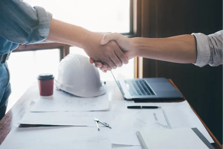 Two people shaking hands over a desk with construction documents, a laptop, coffee cup, and safety helmet, symbolizing agreement in a building project.