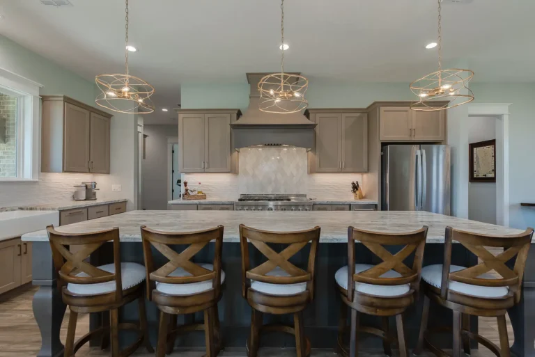 Modern kitchen with a large island and five wooden barstools, pendant lights overhead, gray cabinetry, stainless steel appliances, and a farmhouse sink.