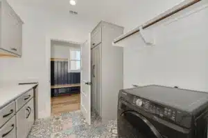 Laundry room with patterned tile, gray cabinets, quartz counters, hanging rod, and black Samsung washer; door opens to mudroom bench.