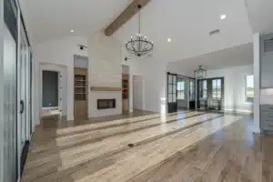 Sunlit open-concept living room with wood-look tile floors, stone fireplace with built-ins, exposed beam, black chandelier, and glass barn doors.