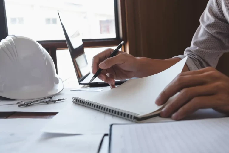 Close-up of a contractor’s hands writing in a notebook beside a laptop, hard hat, and drafting tools—symbolizing permits, inspections, and project planning.