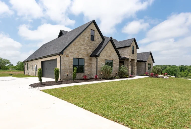 Stone-clad custom home with dark gabled roof, large windows, and a landscaped front yard beside a wide concrete driveway.