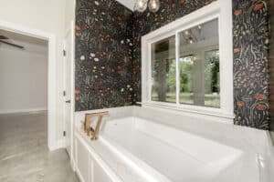 Bathroom accent wall that adds warmth without trend fatigue, featuring dark floral wallpaper behind a white soaking tub with brass fixtures.