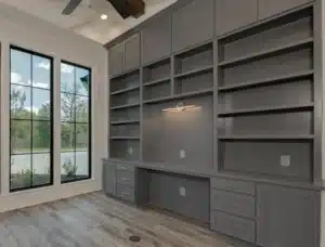 Modern home office with built-in gray shelving, cabinetry, and a long desk, under a ceiling fan beside tall black-framed windows.