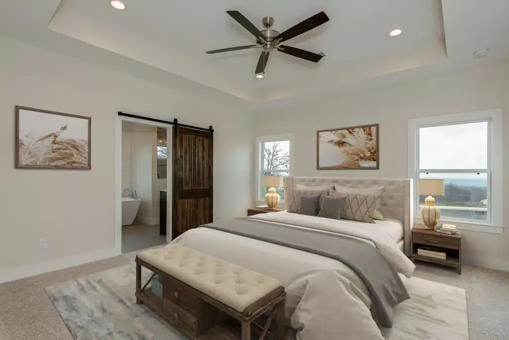 Current image: Serene primary bedroom with upholstered bed, tufted bench, tray ceiling fan, sliding barn door to bath, neutral décor, two windows and lamps.
