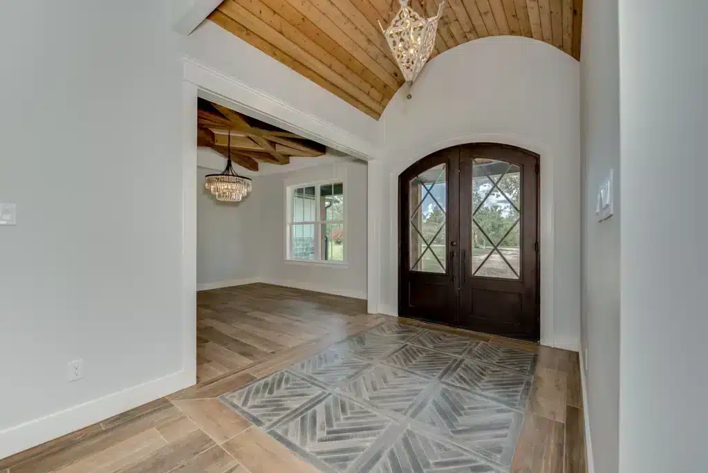Current image: Bright custom home entryway with arched double front doors, wood plank vaulted ceiling, statement chandelier, and clean white trim details.