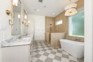 Bathroom with checkered floor, white tub under a window, white granite vanity, beige-yellow walls, and two mirrors over twin sinks—Custom Home Lighting Guide for Every Room.